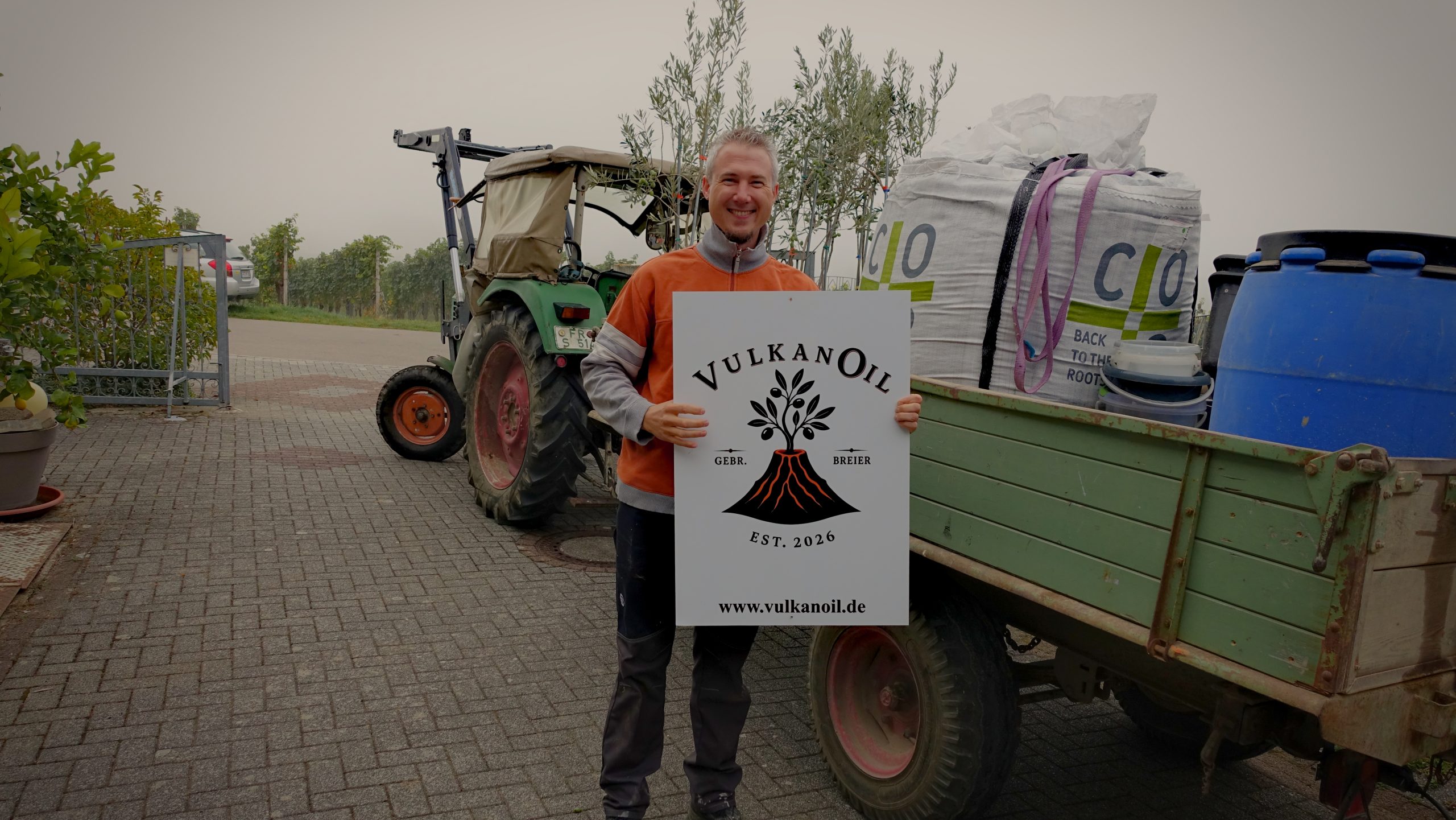 Mann mit dem VulkanOil-Schild vor einem Traktor und jungen Olivenbäumen.