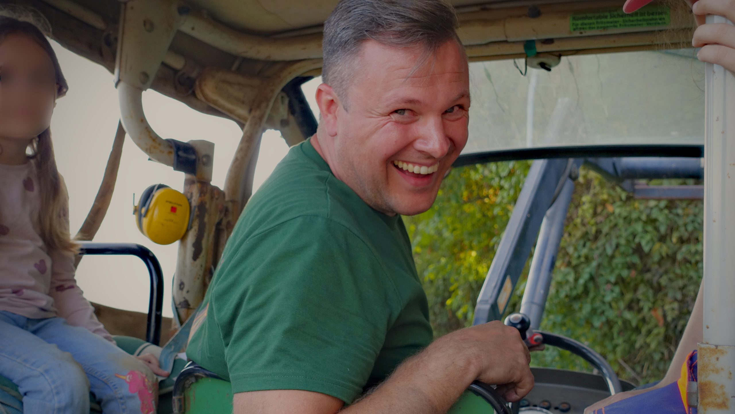 Ein Helfer lächelt auf einem Traktor; das Gesicht des Kindes daneben ist unkenntlich gemacht.